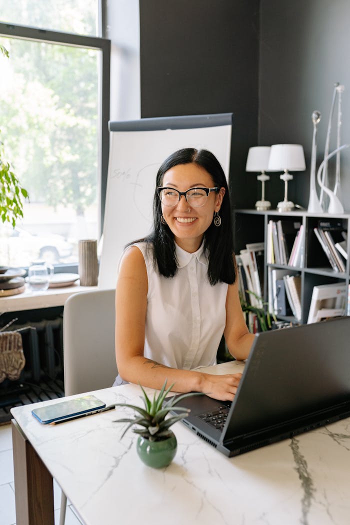 Services Smiling Asian businesswoman using laptop in a stylish, well-lit office with plants.
